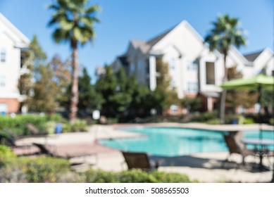 Blurred Image Outdoor Swimming Pool And Woven Rattan Pool Beds With Modern Apartment Houses In Background. Apartment Backyard Landscape With Swimming Pool, Palm Trees And Clear Blue Sky In Texas, US.