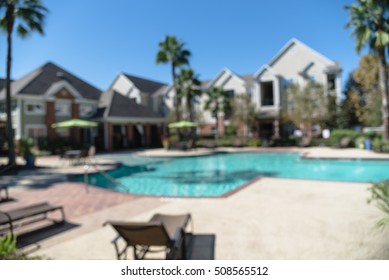 Blurred Image Outdoor Swimming Pool And Woven Rattan Pool Beds With Modern Apartment Houses In Background. Apartment Backyard Landscape With Swimming Pool, Palm Trees And Clear Blue Sky In Texas, US.