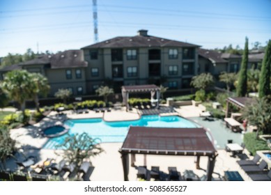 Blurred Image Outdoor Swimming Pool And Woven Rattan Pool Beds With Modern Houses In Background. Elevated View Apartment Backyard Landscape With Swimming Pool, Palm Trees, Clear Blue Sky In Texas, US.