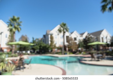 Blurred Image Outdoor Swimming Pool And Woven Rattan Pool Beds With Modern Apartment Houses In Background. Apartment Backyard Landscape With Swimming Pool, Palm Trees And Clear Blue Sky In Texas, US.