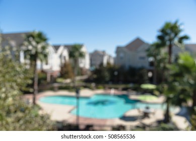 Blurred Image Outdoor Swimming Pool And Woven Rattan Pool Beds With Modern Houses In Background. Elevated View Apartment Backyard Landscape With Swimming Pool, Palm Trees, Clear Blue Sky In Texas, US.