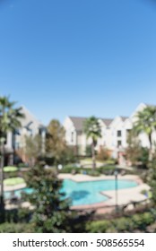 Blurred Image Outdoor Swimming Pool And Woven Rattan Pool Beds With Modern Houses In Background. Elevated View Apartment Backyard Landscape With Swimming Pool, Palm Trees, Clear Blue Sky In Texas, US.