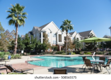 Blurred Image Outdoor Swimming Pool And Woven Rattan Pool Beds With Modern Apartment Houses In Background. Apartment Backyard Landscape With Swimming Pool, Palm Trees And Clear Blue Sky In Texas, US.