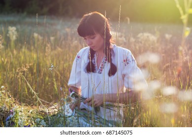 Blurred image of a woman who weaves a wreath of wild flowers in a meadow at sunset. Girl in ancient national clothes. Light effect from the sun's rays