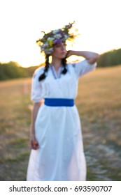 Blurred image of a young woman in a long white embroidered shirt and in a wreath of wild flowers in the field at sunset. Girl in ancient national clothes.