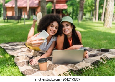 Blurred Interracial Same Sex Couple Taking Selfie Near Laptop And Coffee In Park