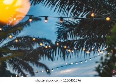 Blurred Light Bokeh With Coconut Palm Tree Background On Sunset, Yellow String Lights With Bokeh Decor In Outdoor Restaurant