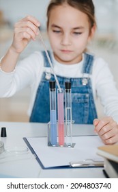 Blurred Little Girl Doing Home Science Project, Filling A Pipette With Liquid From The Flask. Behind A Table.