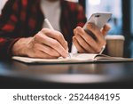 Blurred male in casual outfit holding modern smartphone and making notes in open notepad while sitting at table in cafe