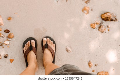 Blurred Man In Black Slippers  Flip Flop Feet Standing On The Beach With A Wave Of Foaming Gentle Beneath Them.Top View