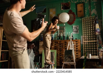 Blurred Man With Vintage Camera Pointing With Hand Near Cheerful African American Woman Standing On Stool With Cup Of Tea