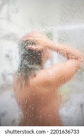 Blurred Middle-aged Unrecognizable Caucasian Woman Enjoying Hot Shower In Steamy Bathroom Behind The Focused Water Splashed Glass Of The Shower Cabin. Selective Focus.