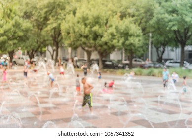 Blurred Motion Kids Playing With Water At Playground In Green Tree Urban Park Fountain. Hot Sunny Day During Summer In Houston, Texas, USA. Water Gushes And Splashes, Sprinkles. Row Of Cars On Street