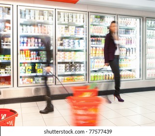 Blurred Motion Of People Walking Near Refrigerator In Shopping Centre With Baskets