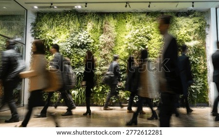 A blurred motion shot captures the dynamic movement of diverse people passing by a vibrant, living green wall in a modern interior space.