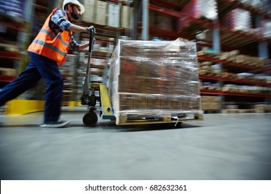 Blurred motion shot of warehouse worker wearing hardhat and reflective jacket pushing moving cart with boxes along isle between tall racks