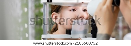 blurred oculist checking eyesight of girl on vision screener in optics shop, banner