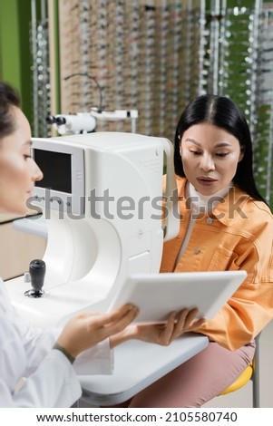 blurred oculist showing digital tablet to asian woman near vision screener in optics salon