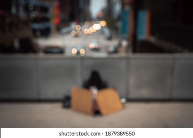 A Blurred Out Of Focus Photograph Of A Homeless Man Wearing A Coat And Hoodie And Sitting On An Overpass Sidewalk In Downtown Chicago With Cup And Cardboard Signs.