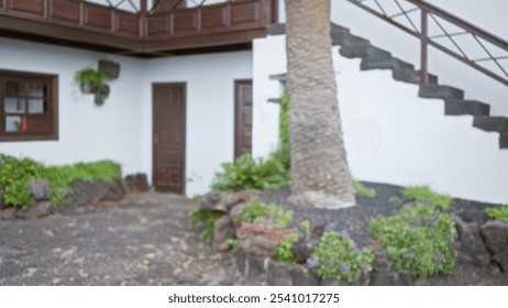 Blurred outdoor scene in lanzarote, spain with defocused background, including a tree, plants, and a traditional white building with wooden doors and a staircase. - Powered by Shutterstock