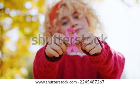 Blurred pensive serious beautiful caucasian blond woman in warm red sweater holding pink ribbon with two hands. Breast and abdominal cancer awareness. High quality photo