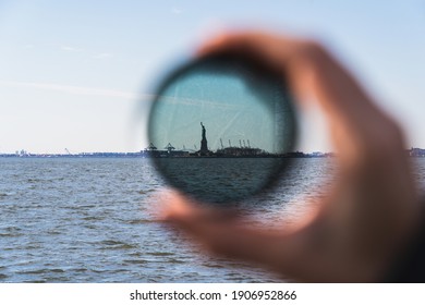 A Blurred Photo Of A Hand Holding Up A Lens Filter, With The Statue Of Liberty In Focus