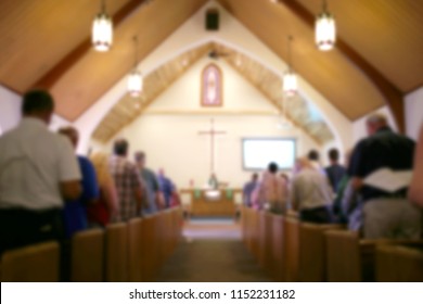 A blurred photo of the inside of a church sanctuary that is filled with people in the pews, and the pastor stands under a large cross at the altar. - Shutterstock ID 1152231182