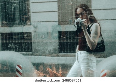 Blurred photo of stylish girl with camera taking selfies in reflection of scratched window on city street. Young woman in white jeans. People outdoors