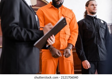 blurred prosecutor writing on clipboard near handcuffed african american man and bailiff in court