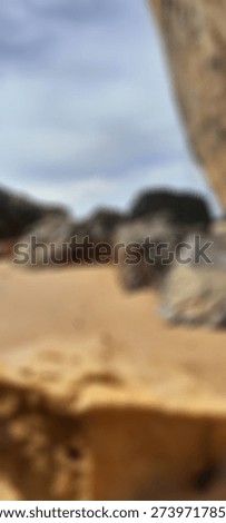 Blurred sandy beach with soft focus rocks and defocused coastal landscape, abstract seaside background featuring warm sand tones and muted cloudy sky