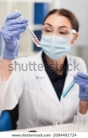 Blurred scientist in medical mask and latex gloves looking at test tube in laboratory