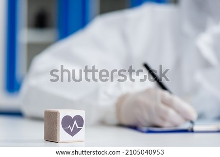 blurred scientist in personal protective equipment writing on clipboard near cube with heart sign