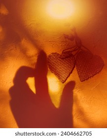 Blurred shadow of female hand and two wooden heart on rough yellow wall background. Sunlight and  crossed lines shadows, Minimal aesthetics. Abstract, play of light and shadow