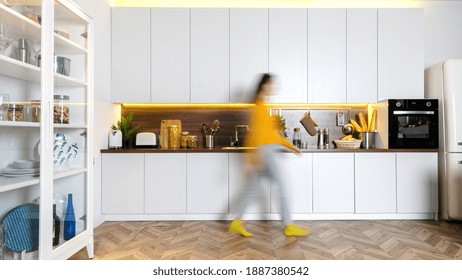 Blurred silhouette of young woman walking at home. Panoramic view of modern interior with white kitchen cupboards, wooden countertop, electric oven - Powered by Shutterstock