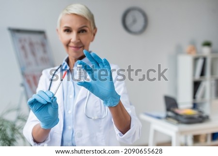 blurred and smiling doctor in latex gloves holding bottle with coronavirus vaccine and syringe in hands