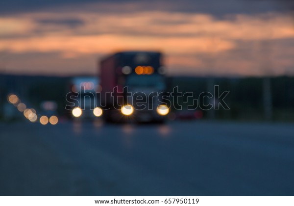 Blurred texture Traffic trucks\
with glowing lights on the highway after sunset. Lights of\
searchlights. Route, road, transport. Blue and orange bright sky at\
dusk.