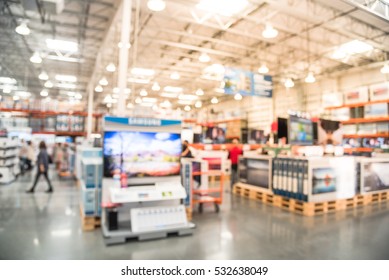 Blurred TVs Shopping At Large Wholesale Club. Television Retail Shop, Row Of Big Screen, Smart TVs Display On Shelves. Customers Browsing To Select And Use Flatbed Cart To Carry TV To Checkout Counter