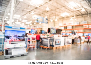 Blurred TVs Shopping Large Wholesale Club. Television Retail Shop, Row Of Big Screen, Smart TVs Display On Shelves. Store Staff Help Customers Select And Use Flatbed Cart Carry TV To Checkout Counter.