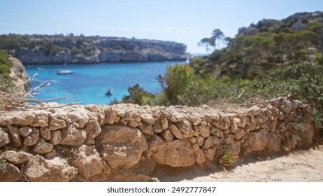 Blurred view of a beautiful rocky coastline in mallorca, spain, framed by a rustic stone wall with vibrant trees and a clear blue sky in the background - Powered by Shutterstock