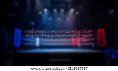 A blurred view of a boxing ring with red and blue corners illuminated by overhead stage lights in a dark stadium setting.