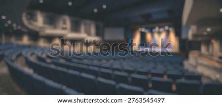 A blurred view of an empty auditorium featuring a brightly lit stage with spotlights and numerous rows of dark seating, ready for an event.