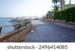 Blurred view of an outdoor coastal walkway with defocused background, railing, path, ocean, and trees on a sunny day.