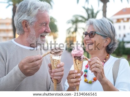 Blurred view of smiling retired couple having fun eating ice cream cone in the park. Focus on ice cream. Joyful elderly people looking into each other eyes laughing