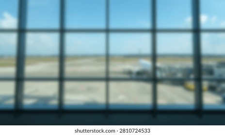 Blurred view through large grid windows of an airport terminal showing the tarmac, runway, and parked airplanes under a clear blue sky.