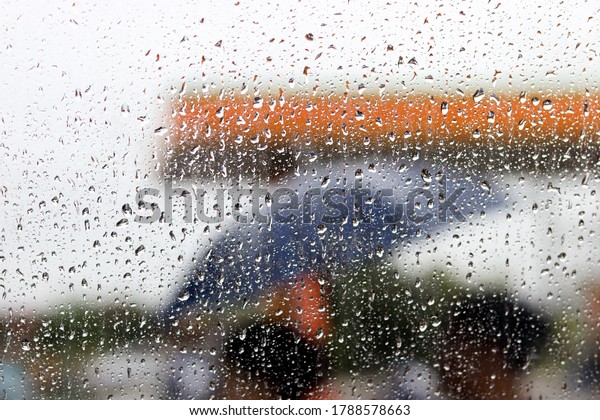 Blurred view of windows cars in gas station\
through car windshield covered in rain drops, Blurred two people\
holding umbrella walk through car\
window.