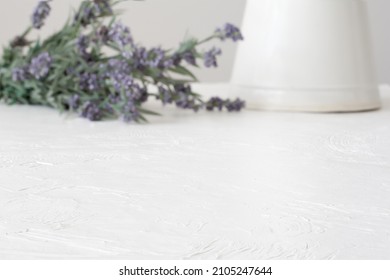 Blurred, Vintage Milk Jug Laying Next To A Bunch Of Lavender On A Textured, White Table. In Focus Foreground For Placing A Digital Product Mockup Or Overlaying With Text.