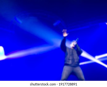 Blurred Vocalist Man, Singer Holding Microphone And Singing A Song On Stage With Blue Light On Dark Background. Blurry Music Performance In Rock Band Concert.