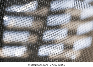 Blurred white objects behind dark wire mesh screen creating abstract texture and mysterious mood, grid pattern covering soft focus background shapes representing protection and obscured vision