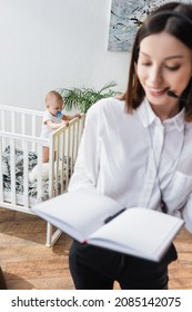 Blurred Woman In Headset Holding Empty Notebook While Working Near Son In Crib At Home