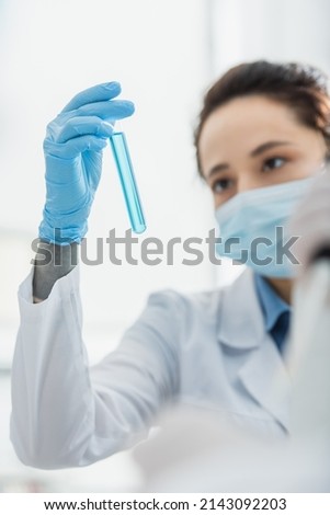blurred woman in medical mask looking at test tube with blue liquid